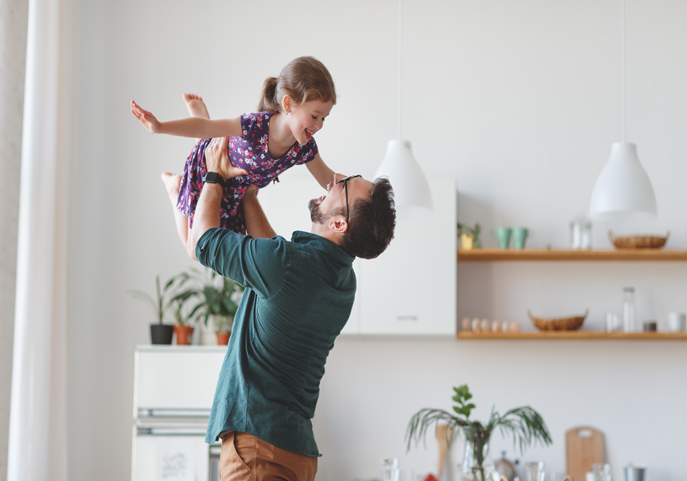 Man and girl keeping spirits high in their apartment home as he lifts her up into an airplane pose.