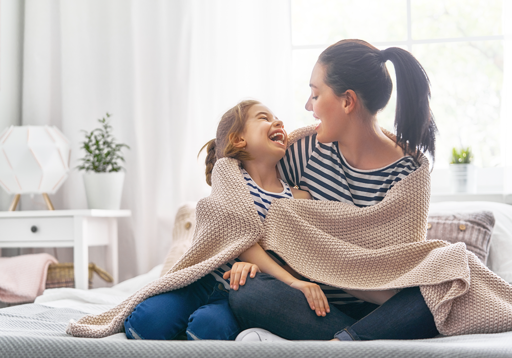 High-spirited mother and daughter wrapped in a blanket on bed in their apartment home bedroom.