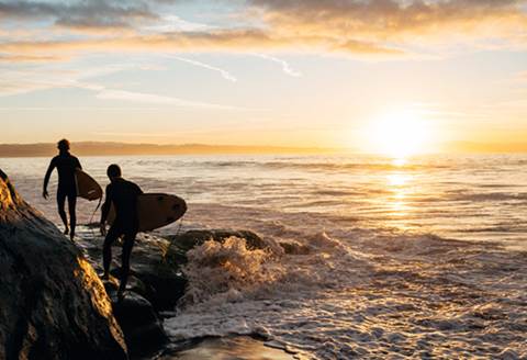 Two surfers walking alongside edge of water at sunset.